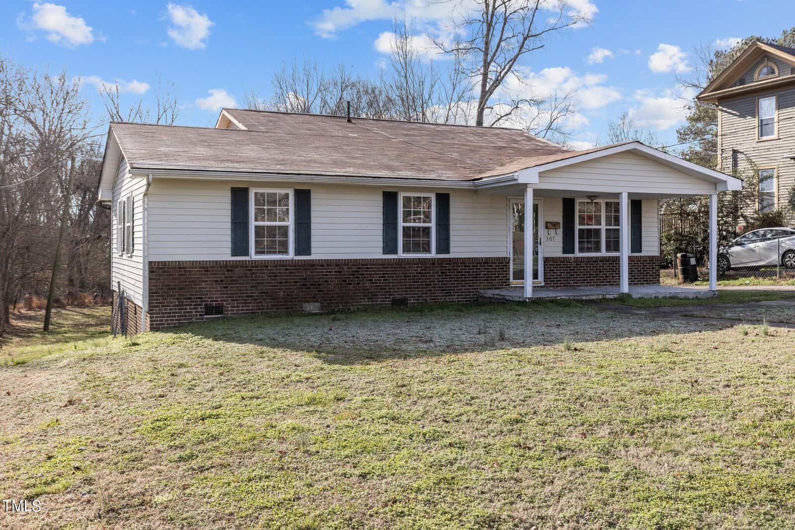 A small, single-story house with a combination of brick and white siding exterior. It features a covered porch with two columns and has two windows flanking the front door. The yard is grassy, and there are other houses and trees in the background.