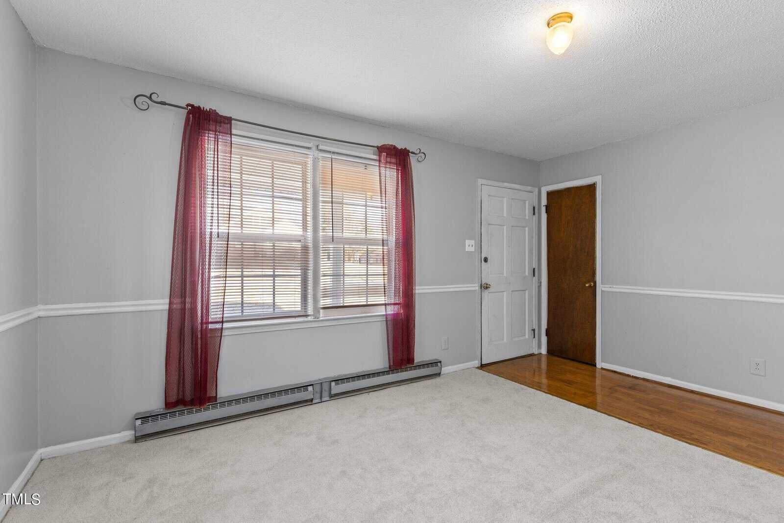 A small, empty room with gray walls and a wooden floor partially covered by a light-colored carpet. A window with red curtains and blinds lets in natural light. Two doors, one wooden and one white, are on opposite walls.