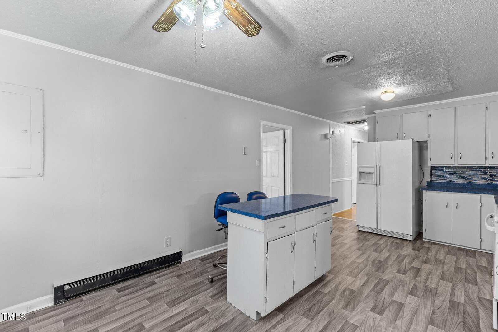 A kitchen with a white fridge, blue countertop island with two blue chairs, and white cabinets. The floor features gray patterned tiles, and a ceiling fan with wooden blades is visible. Walls are a light gray color.
