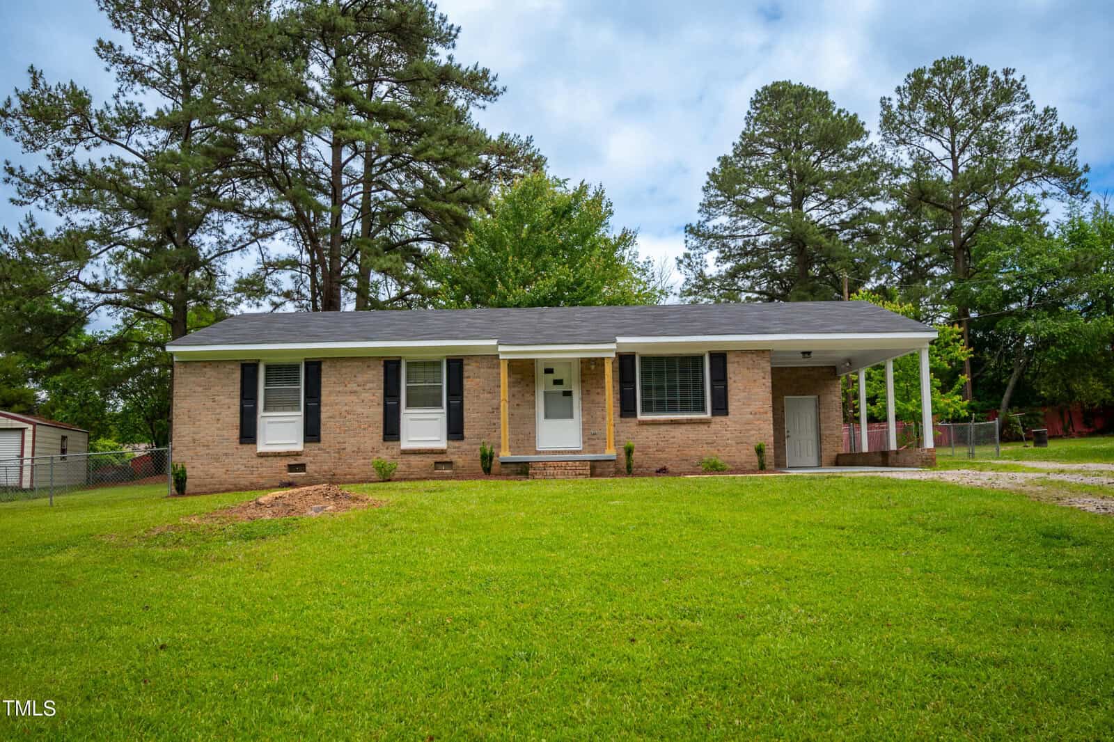 Located at 434 Lightwood Lane in Henderson, NC, this charming brick single-story house features white trim, four front windows, and a small front porch with a white door. A carport is attached to the right. The house is nestled among tall pine trees and surrounded by a grassy lawn under a partly cloudy sky.