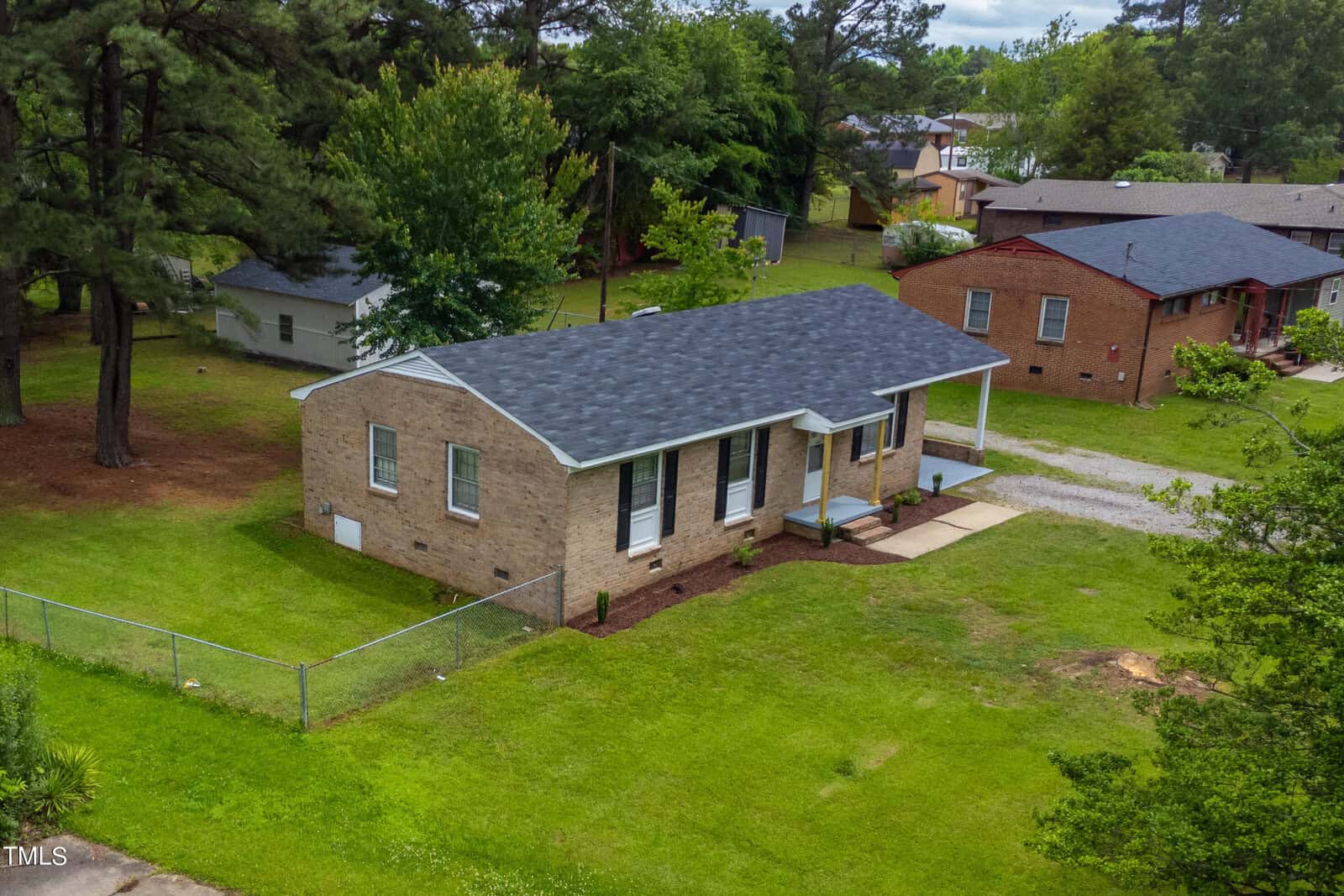 An aerial view of a single-story brick house with a gray shingle roof, a front porch, and a small front yard. The house is surrounded by green grass and trees, with neighboring homes visible in the background. The property is partially enclosed by a chain-link fence.