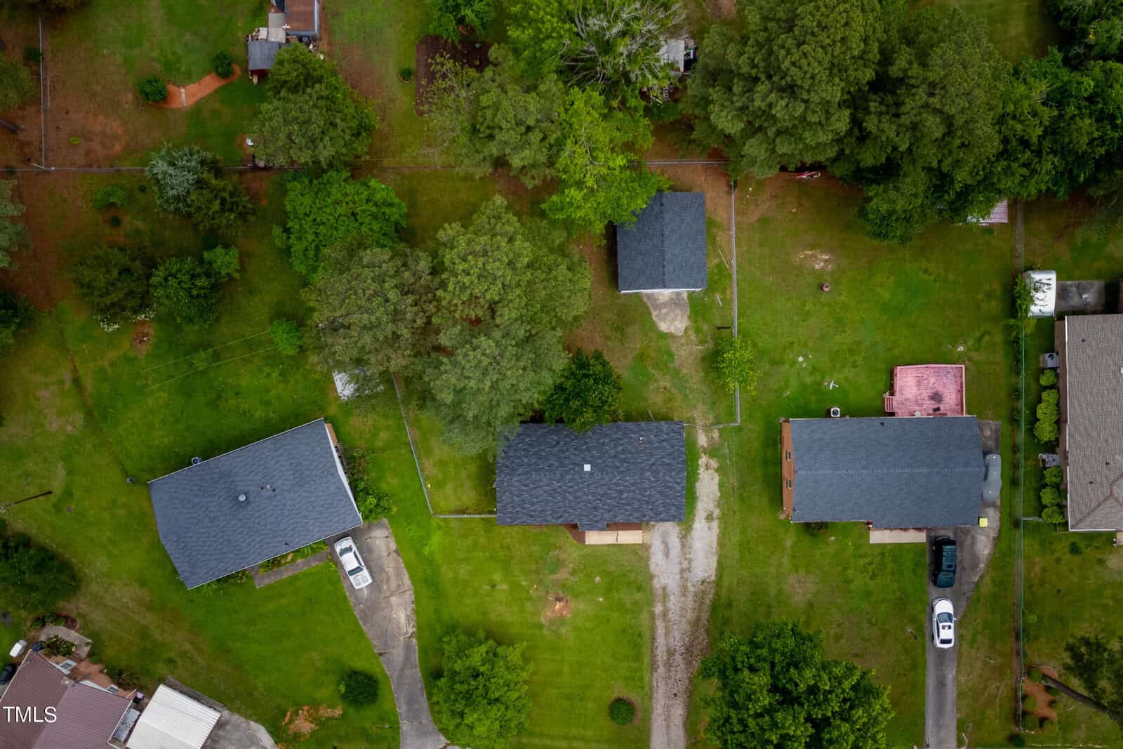 Aerial view of a suburban neighborhood featuring several houses surrounded by green lawns and trees. Two houses have driveways with parked vehicles, and the area appears to be well-maintained with neatly trimmed lawns.