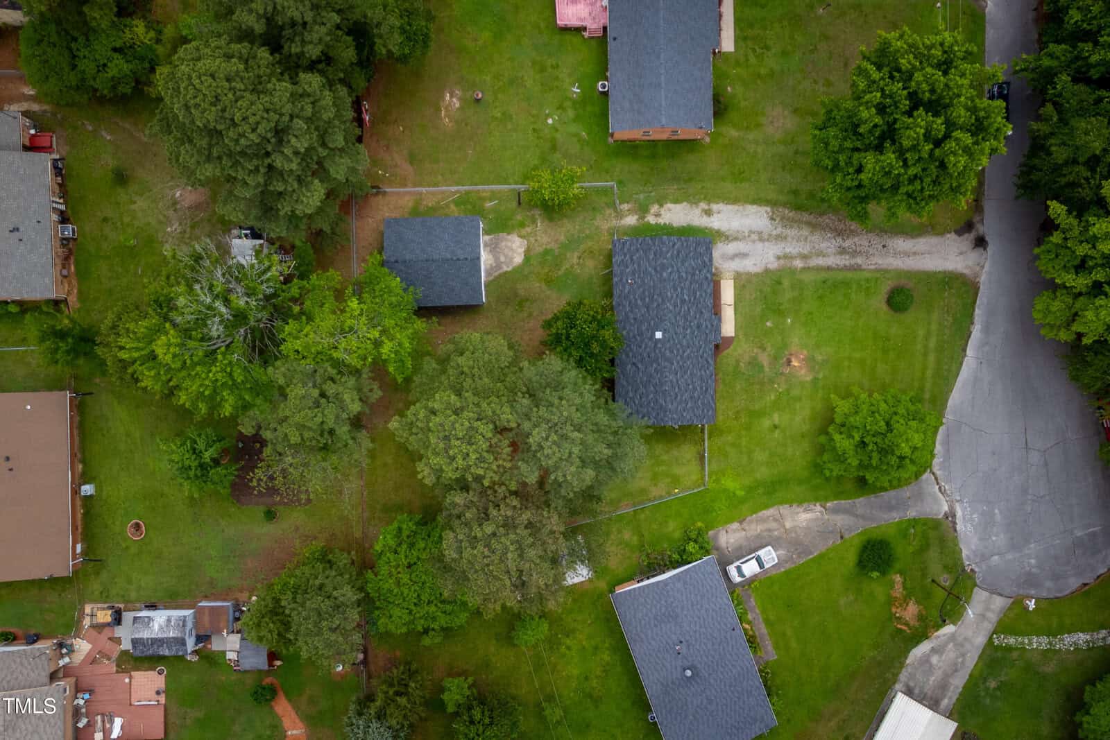 Aerial view of a suburban neighborhood with several single-story homes, lush green lawns, and mature trees. A curved street runs through the area, connecting to driveways and small pathways. One corner shows a parked white vehicle.