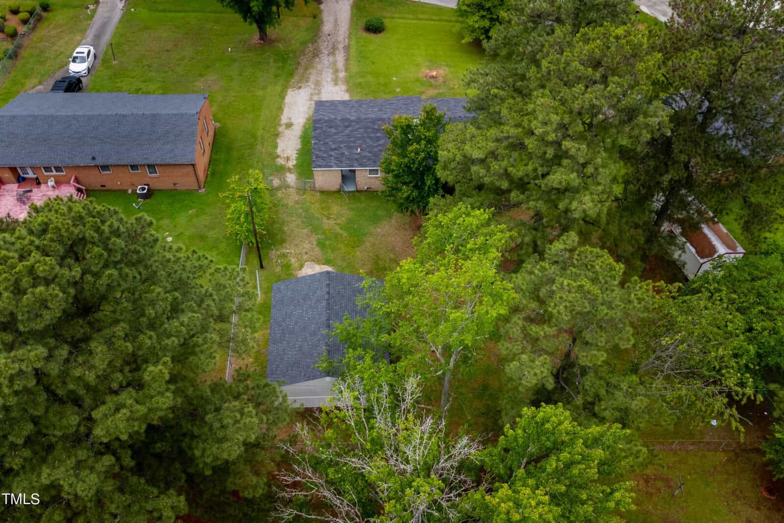 Aerial view of a small neighborhood with several houses surrounded by trees and greenery. A dirt path leads to one of the houses, and a paved road is visible on the left side of the image. The houses have dark shingle roofs and ample outdoor space.
