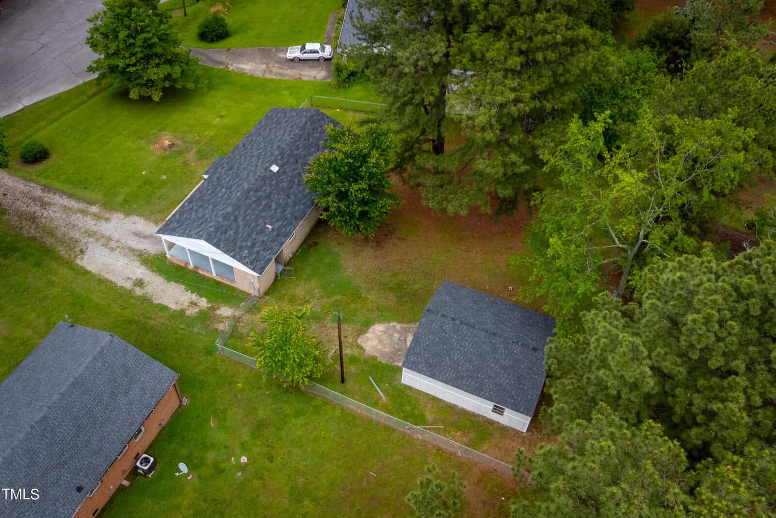 Aerial view of a residential area featuring a house with a gray roof, a driveway, a detached garage, and a yard. Surrounding the property are trees and some greenery. Another house with a gray roof is partially visible at the bottom left of the image.
