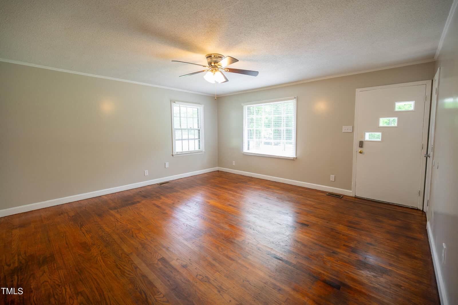A spacious, unfurnished living room with wooden floors, light-colored walls, and a ceiling fan with a light fixture. Two large windows let in natural light, and a white front door with a small glass window is on the right wall.