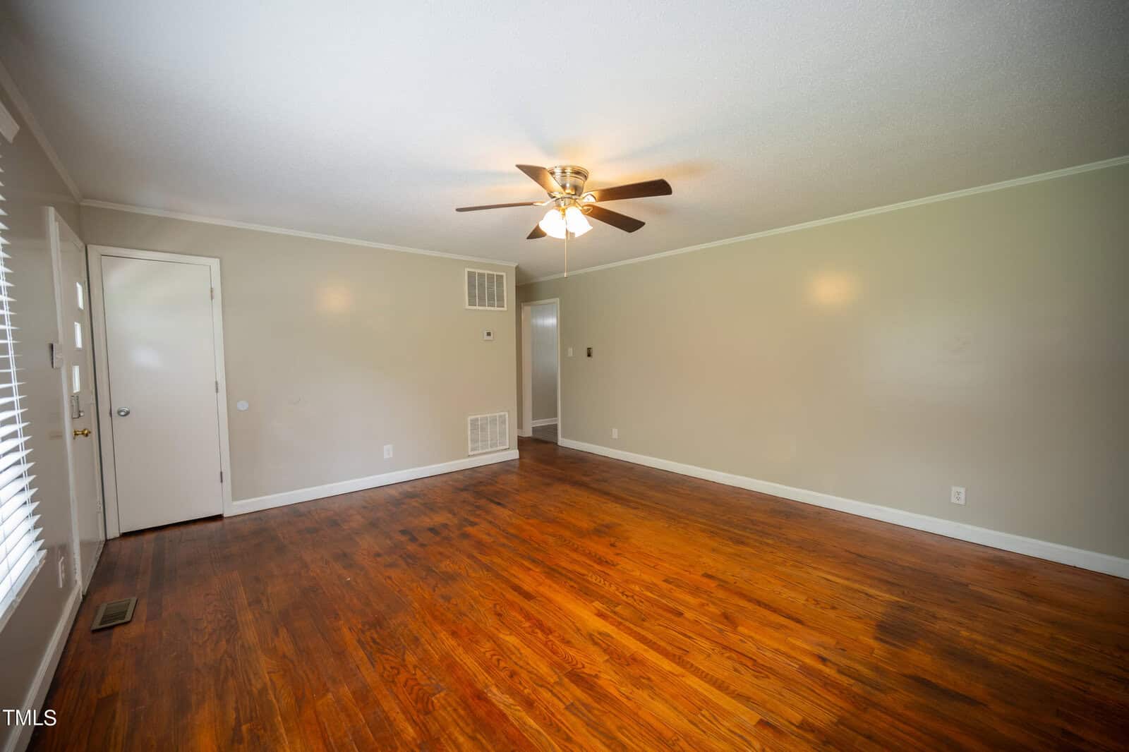 An empty room with light-colored walls and wooden flooring. A ceiling fan with lights is centered on the ceiling. There are closed doors on one side and a partially open door leading to another room. Sunlight filters in through the window with blinds.