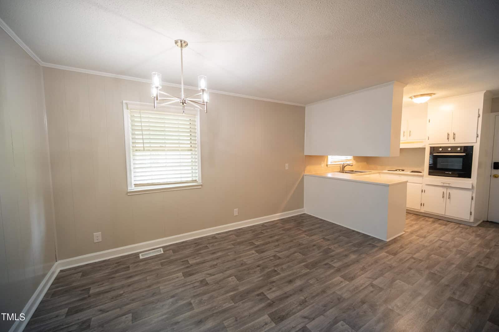 A bright, empty room with light gray walls and wood-look laminate flooring. The room features a window with closed blinds, a modern light fixture on the ceiling, and an adjacent white kitchen with a half wall separating the spaces.