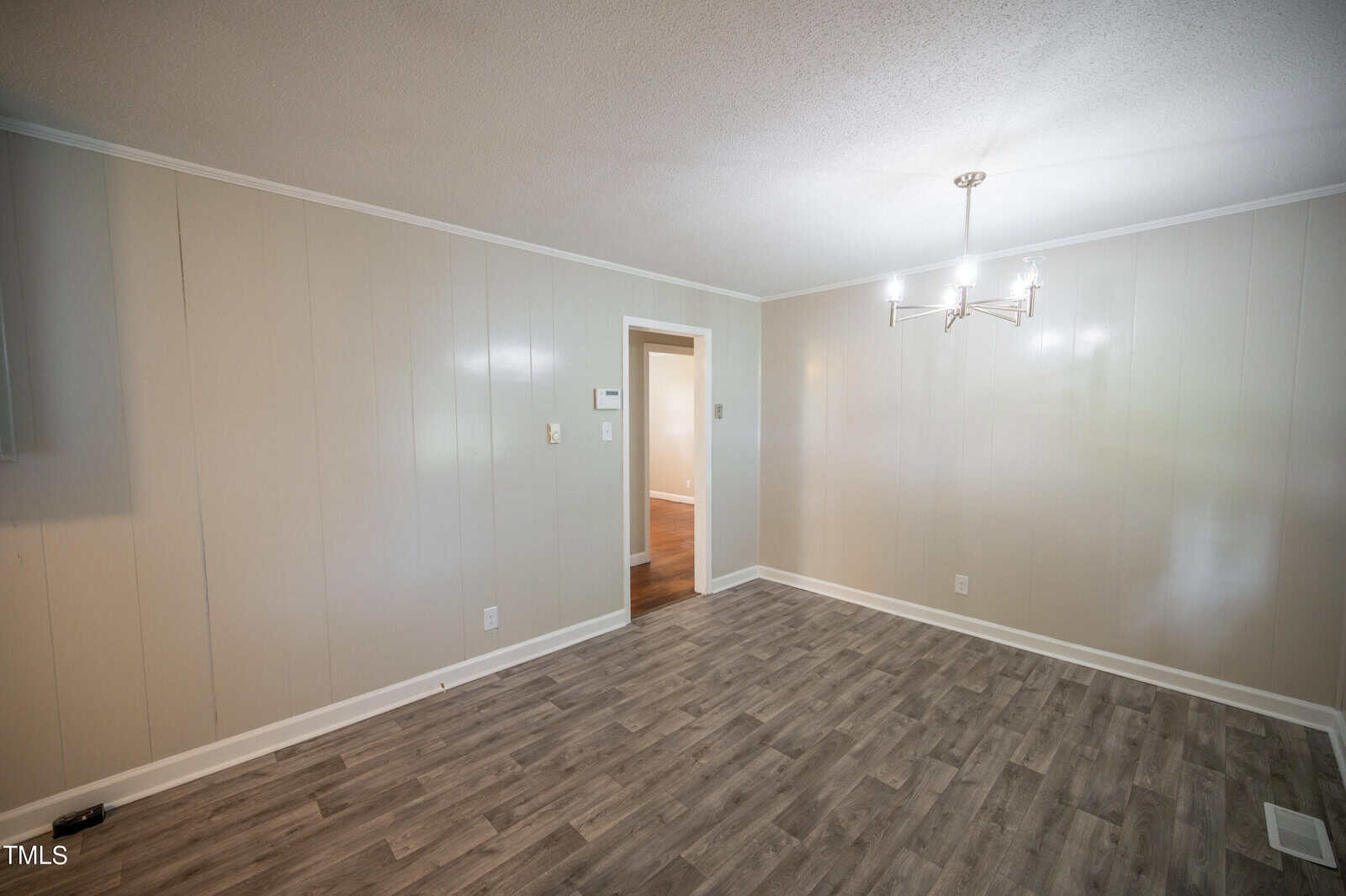 A vacant room with light gray paneled walls, a modern chandelier, and gray wood flooring. There is an open doorway leading to another room with wooden floors visible. The space is well-lit with natural light coming from the unseen windows.