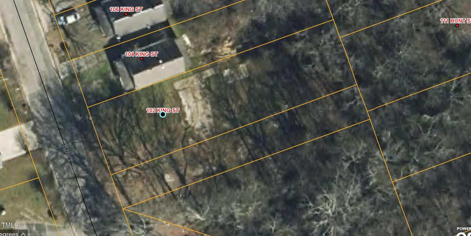 Aerial view of a neighborhood showing houses, trees, and streets. Yellow lines outline property boundaries, and street names are labeled in red. The area is surrounded by dense trees and a road on the left side.