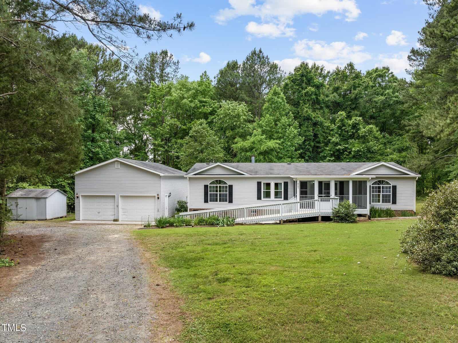 A single-story white house with a gray roof, surrounded by lush green trees. It features a long ramp leading to the entrance and a detached two-car garage to the left. A well-maintained lawn and gravel driveway complete the scene.