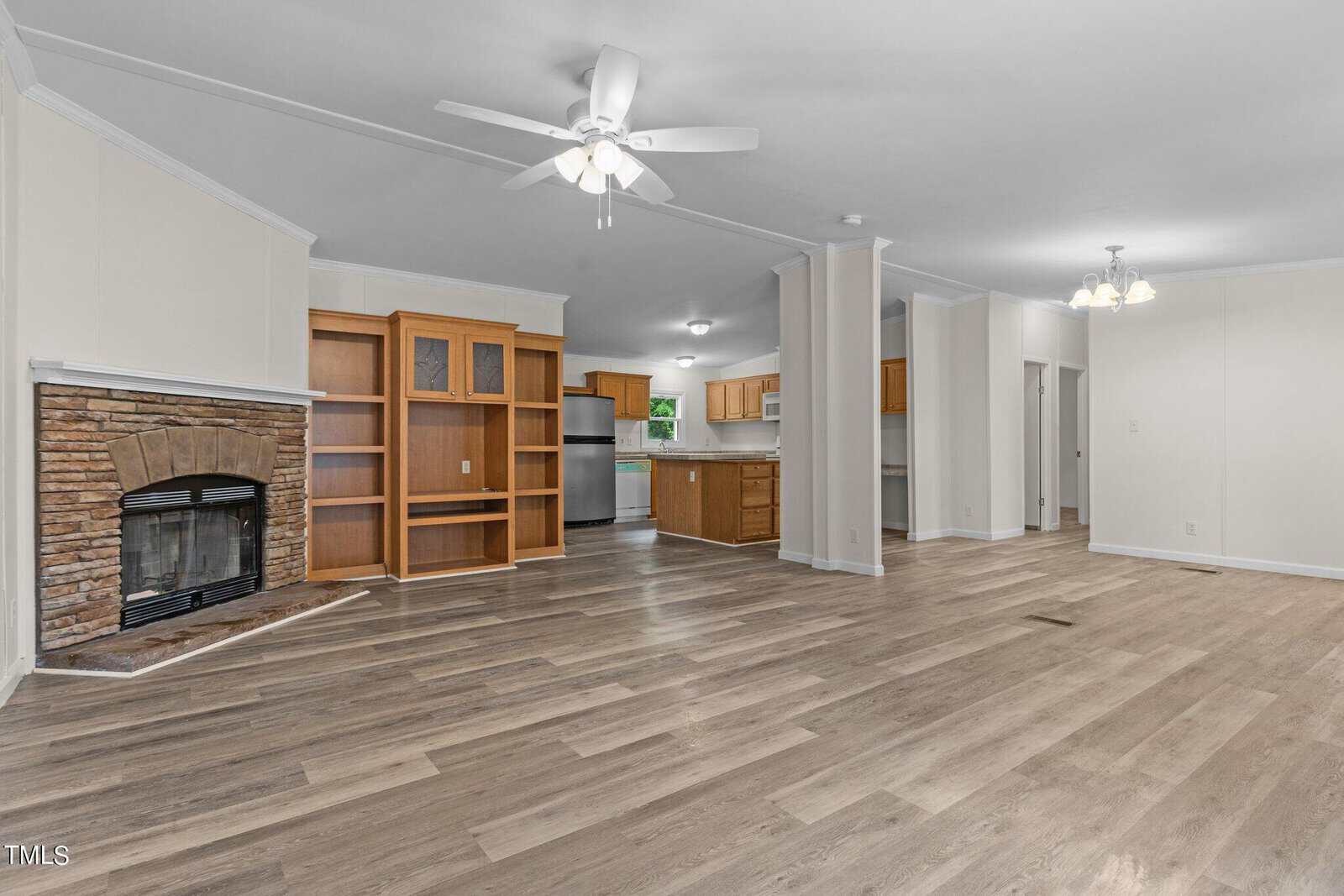 Spacious, open-concept living room with light wood floors, a stone fireplace, ceiling fan, and built-in shelving. The modern kitchen features stainless steel appliances and wood cabinets. Walls are painted in a soft cream color.