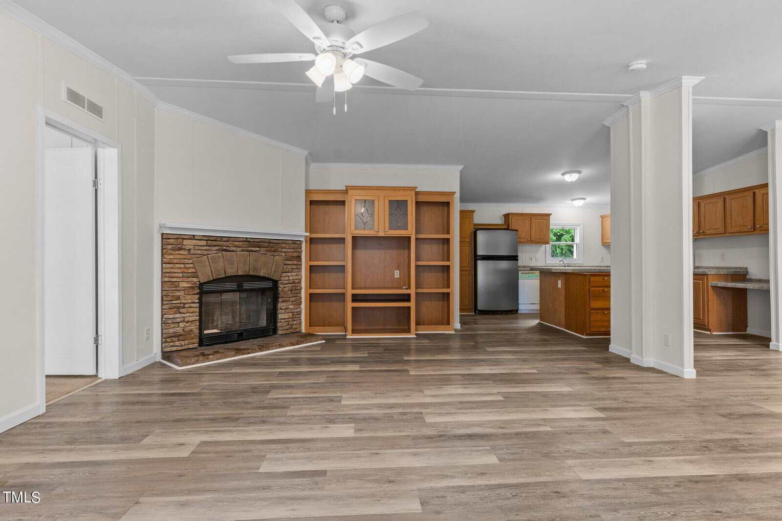 Spacious open living area with wood flooring, featuring a built-in wooden cabinet, stone fireplace, and ceiling fan. Kitchen with stainless steel appliances and wooden cabinets is visible in the background. Walls are painted in neutral tones.