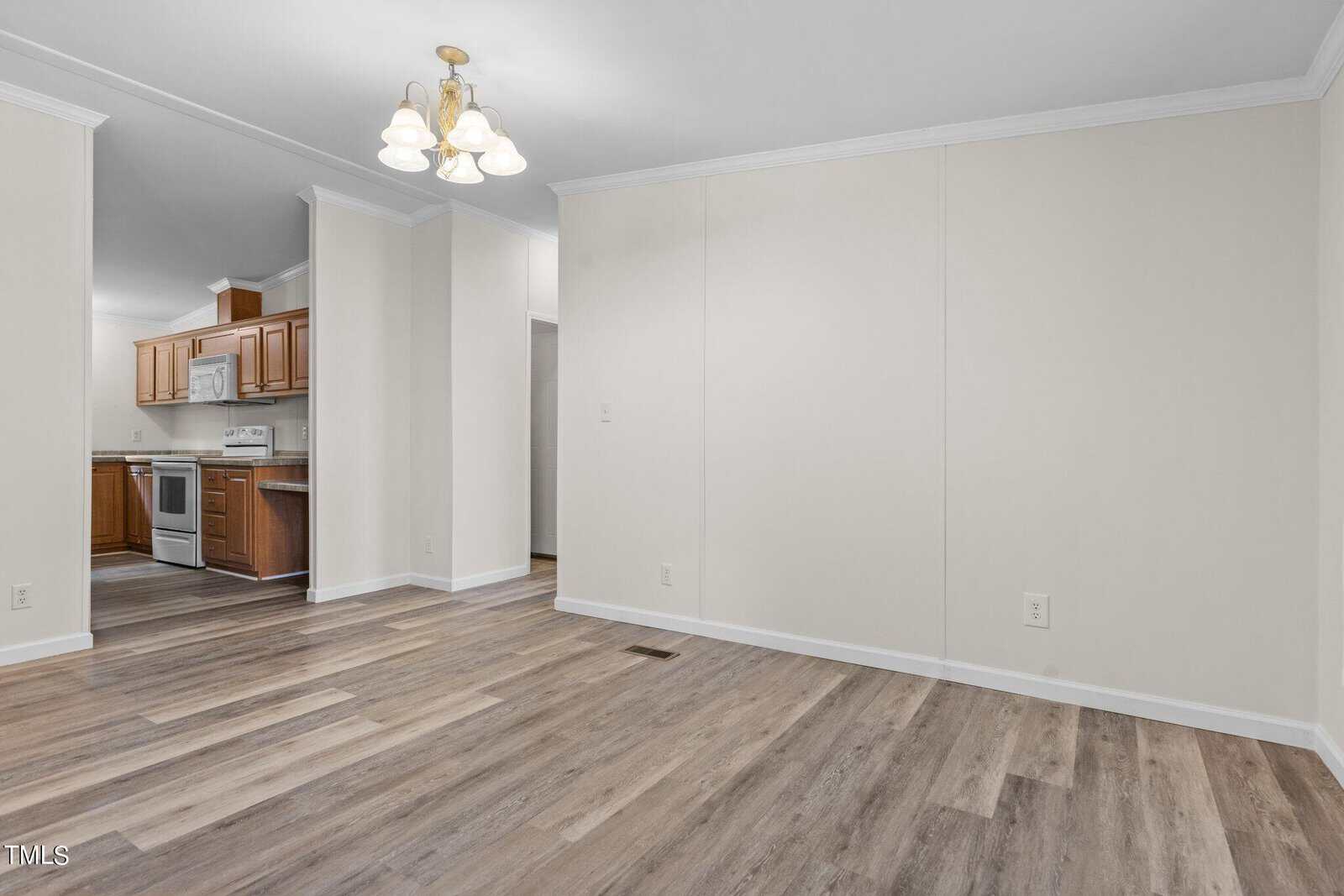 An empty room with light-colored walls and wood-patterned flooring. A doorway leads to a kitchen with wooden cabinets and white appliances. A ceiling light fixture with three bulbs hangs from the ceiling.