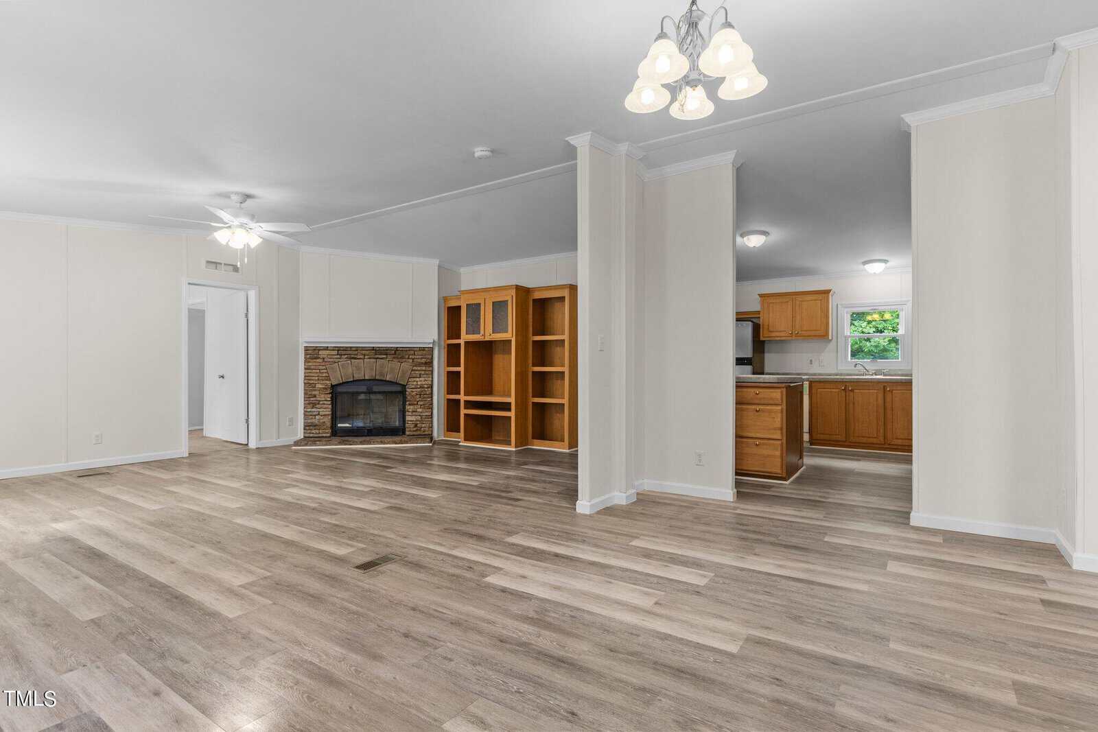 Spacious open-concept living area with light wood flooring, ceiling fan, and chandelier. Features a stone fireplace and built-in shelves, leading to a kitchen with wooden cabinets and a small window. Walls are painted in a light neutral color.