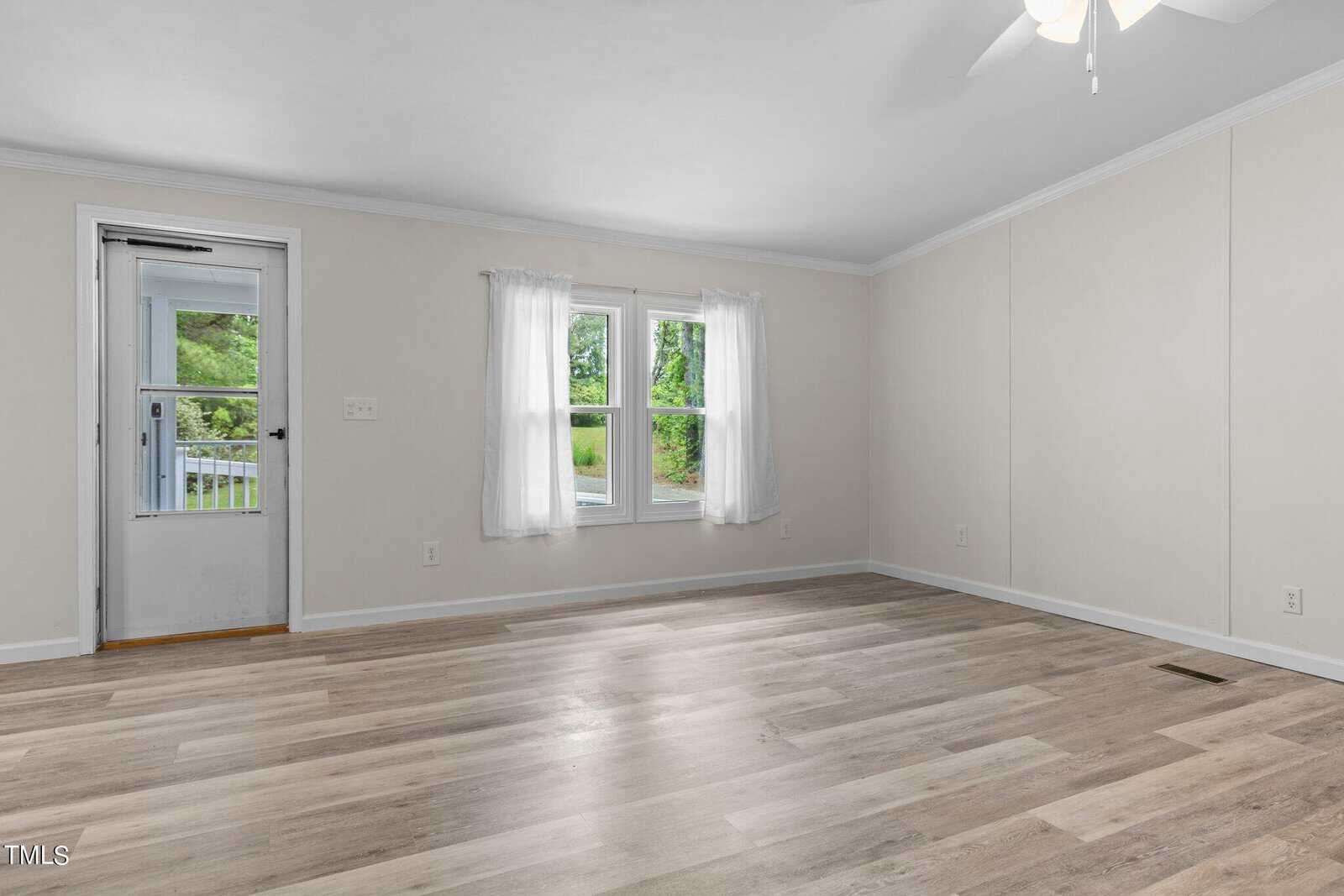 A bright, empty room with light wood flooring, cream-colored walls, and a ceiling fan. A window with sheer curtains and a glass door open onto a green view outside.