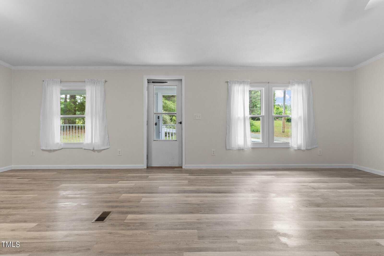 An empty room with light-colored walls and wooden flooring. Two windows on the right wall and one by a door let in natural light. The windows have white curtains, and the view outside shows greenery.