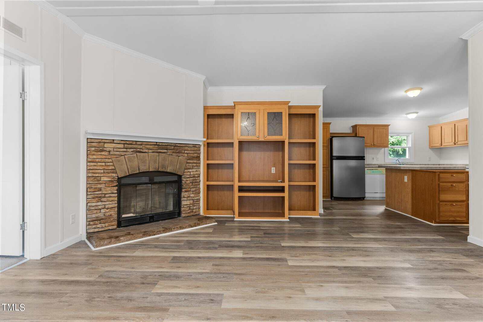 Spacious living room with light wood floors, a stone fireplace on the left, and a built-in wooden bookshelf. The room opens into a kitchen with wooden cabinets, a stainless steel refrigerator, and a dining area.
