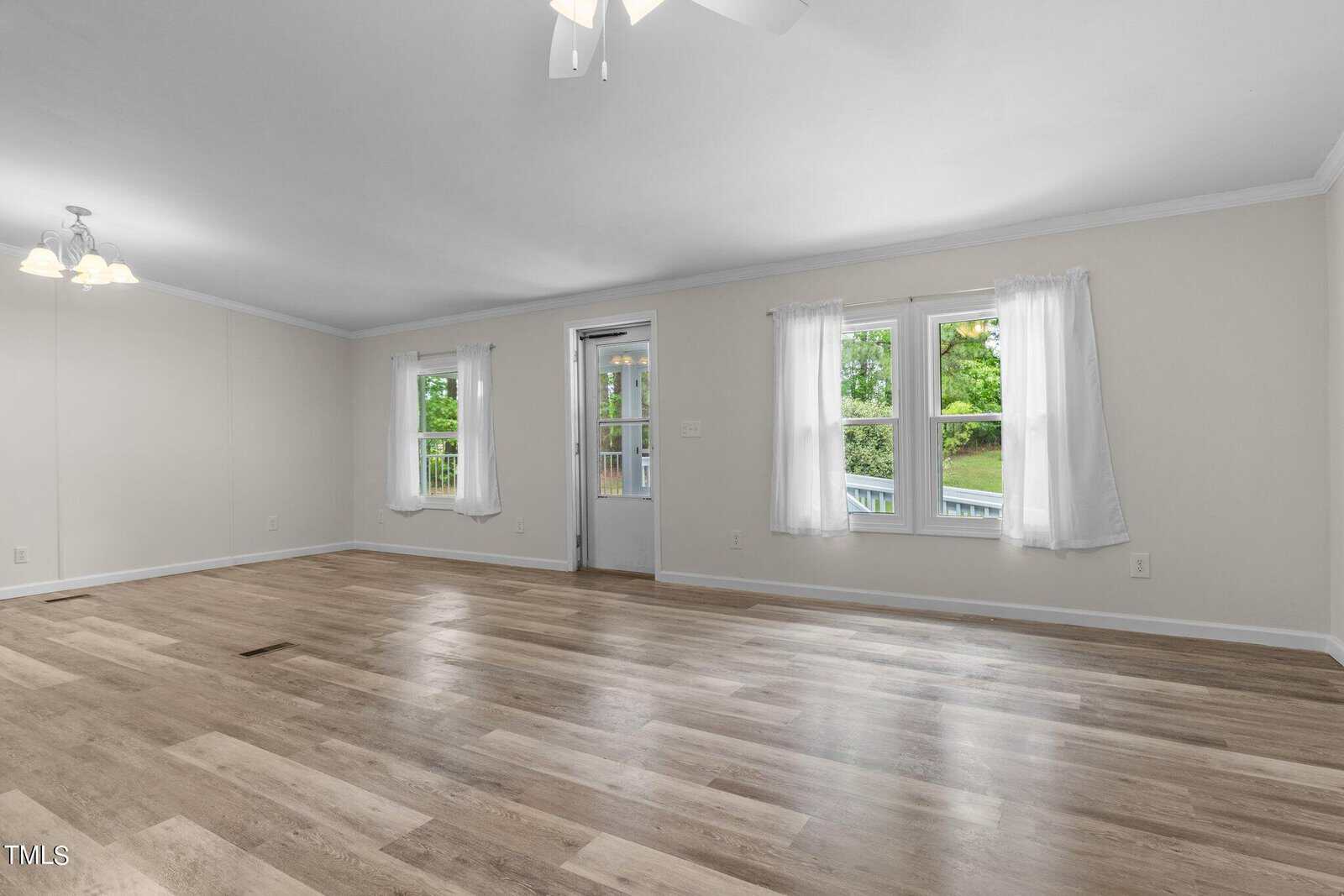 A spacious, empty living room with light wood flooring, white walls, and large windows covered with sheer white curtains. A ceiling fan with lights is on the white ceiling, and a central door with glass panels leads outside.