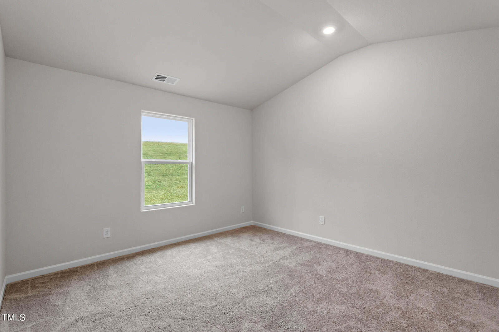 An empty room with beige carpet and light gray walls. A single window shows a grassy field outside. The ceiling is vaulted with a small recessed light.