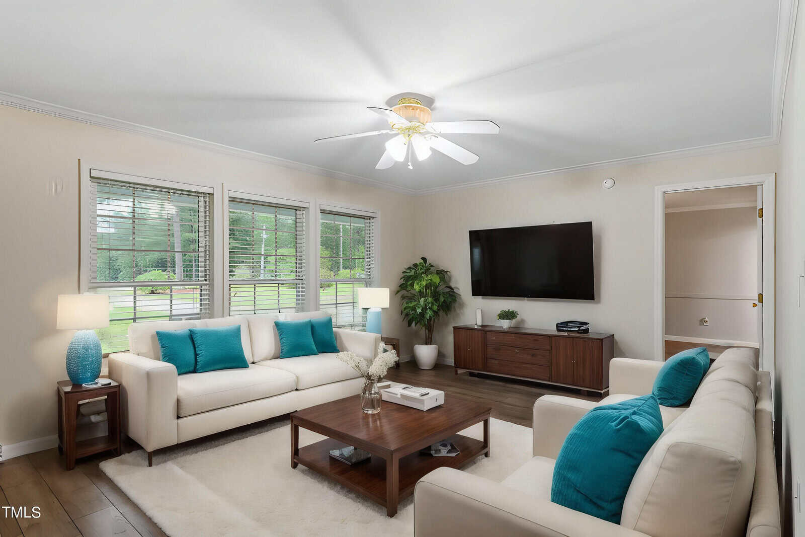 A cozy living room with a ceiling fan and three white sofas accented with blue pillows. A wooden coffee table sits on a white rug. A flat-screen TV is mounted on the wall above a wooden cabinet. Large windows let in natural light.