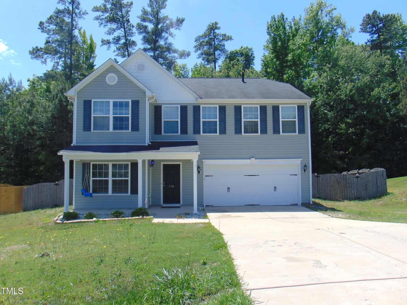 Located at 716 Pitch Pine Drive in Creedmoor, this two-story house boasts blue-gray siding and black shutters, complemented by a charming covered porch and a spacious double garage. The grassy front lawn and towering trees complete the picturesque setting under a clear blue sky.