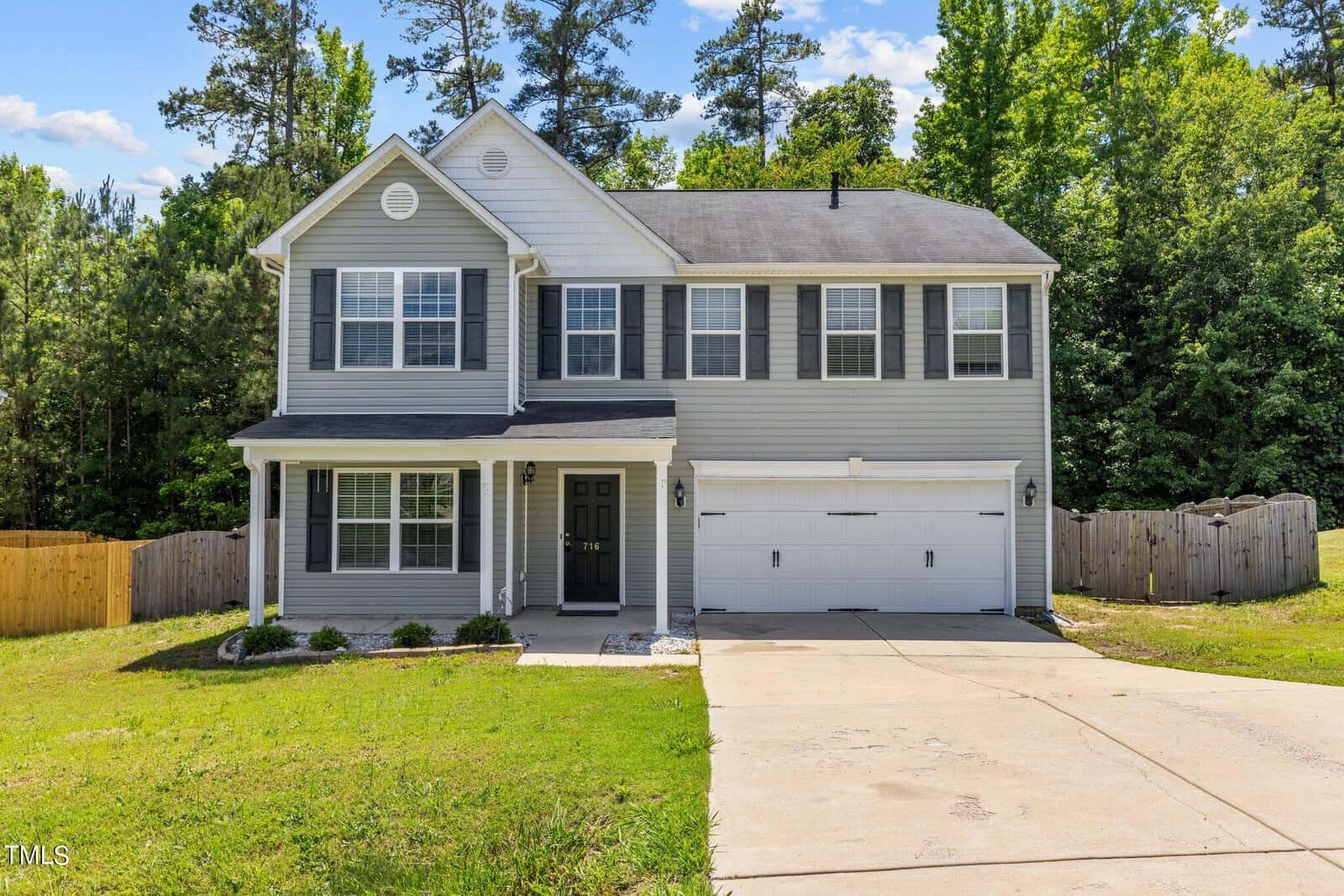 A two-story gray house with white trim is shown, featuring a covered front porch and a two-car garage. It is set against a backdrop of tall trees under a blue sky. The driveway and front yard are visible in the foreground.