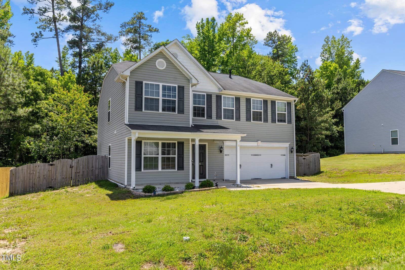 A two-story gray house with white trim and a small front porch. It has a two-car garage, a neatly maintained lawn, and is surrounded by lush green trees and a wooden fence on one side. A clear blue sky is overhead.