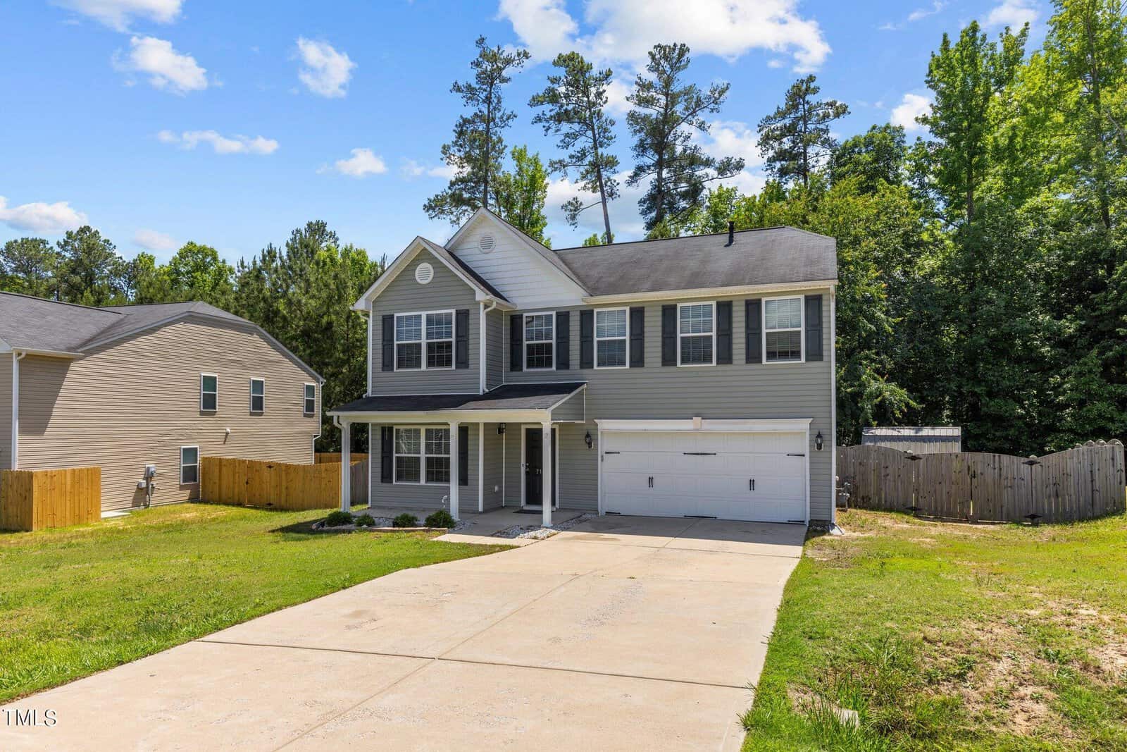 A two-story suburban house with gray siding, white trim, and a black roof, located in a residential area. The front yard features a well-maintained lawn and a paved driveway leading to a two-car garage. Tall trees are visible in the background.