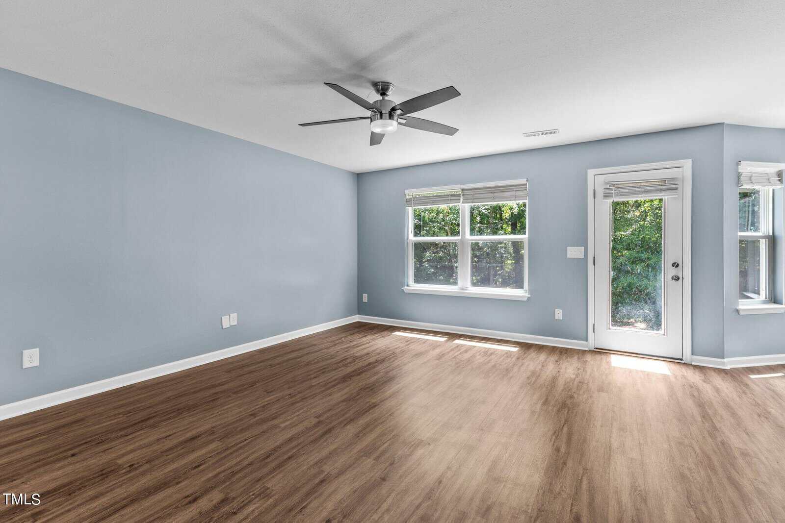 A spacious, empty room with light blue walls and wood flooring. It features a ceiling fan and large windows filling the space with natural light. A glass door opens to an outdoor area with greenery visible through the window.
