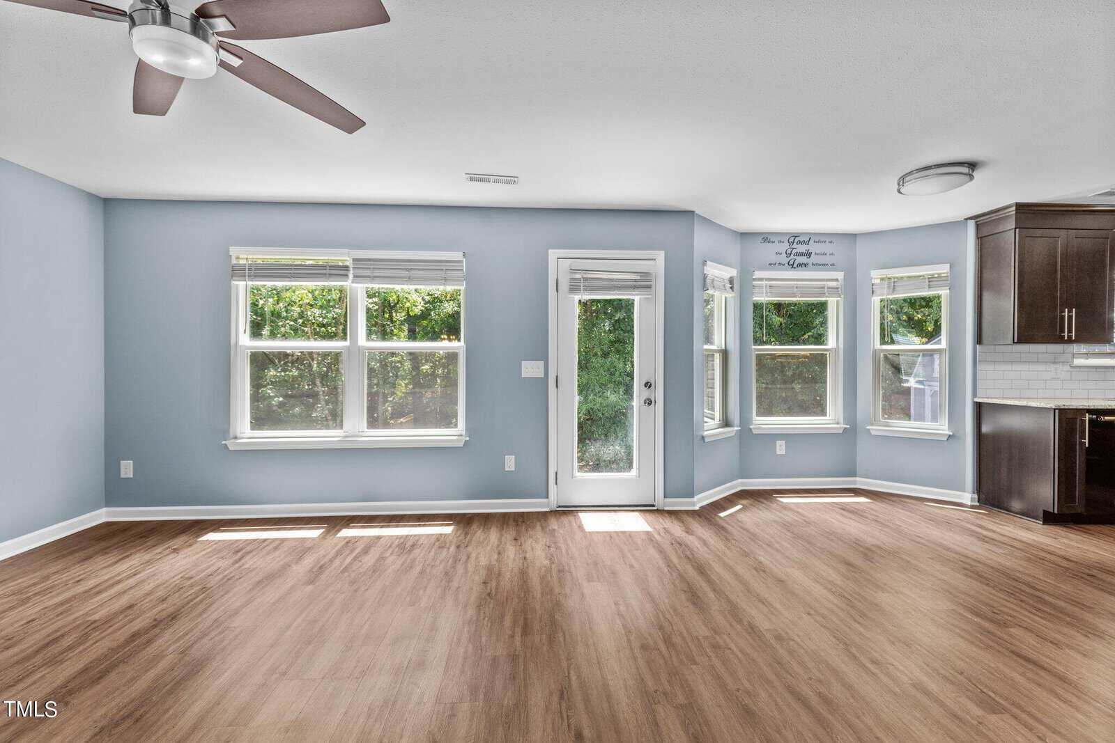 An empty room with light blue walls and wooden floors. It features three large windows, a glass door, and a ceiling fan. There is a partial view of a kitchen with dark cabinets to the right. Sunlight streams in, creating a bright atmosphere.