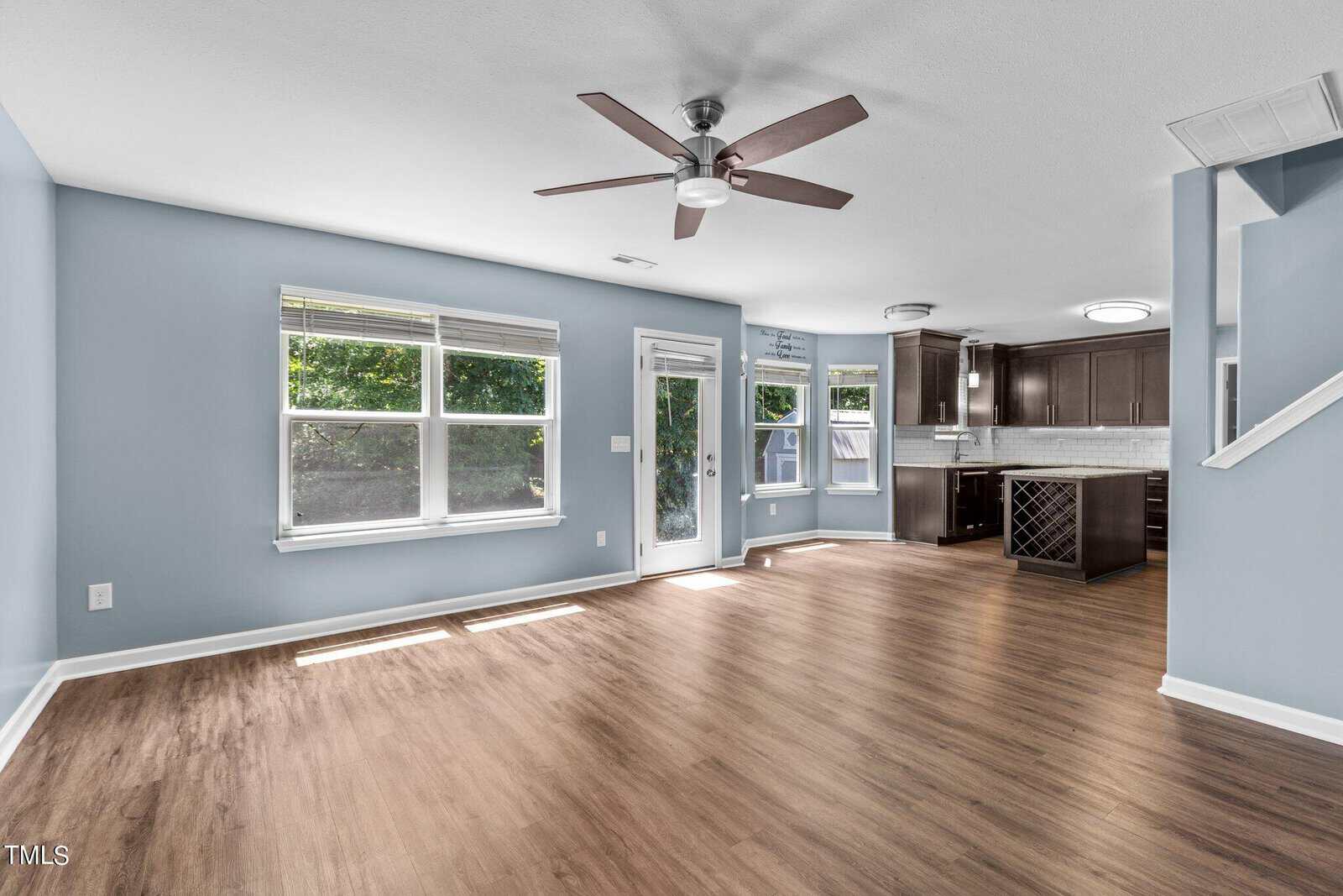 Spacious living area with light blue walls and wooden flooring. Ceiling fan above, large windows and a glass door leading to an outdoor area. Open kitchen with dark cabinets and an island in the background.