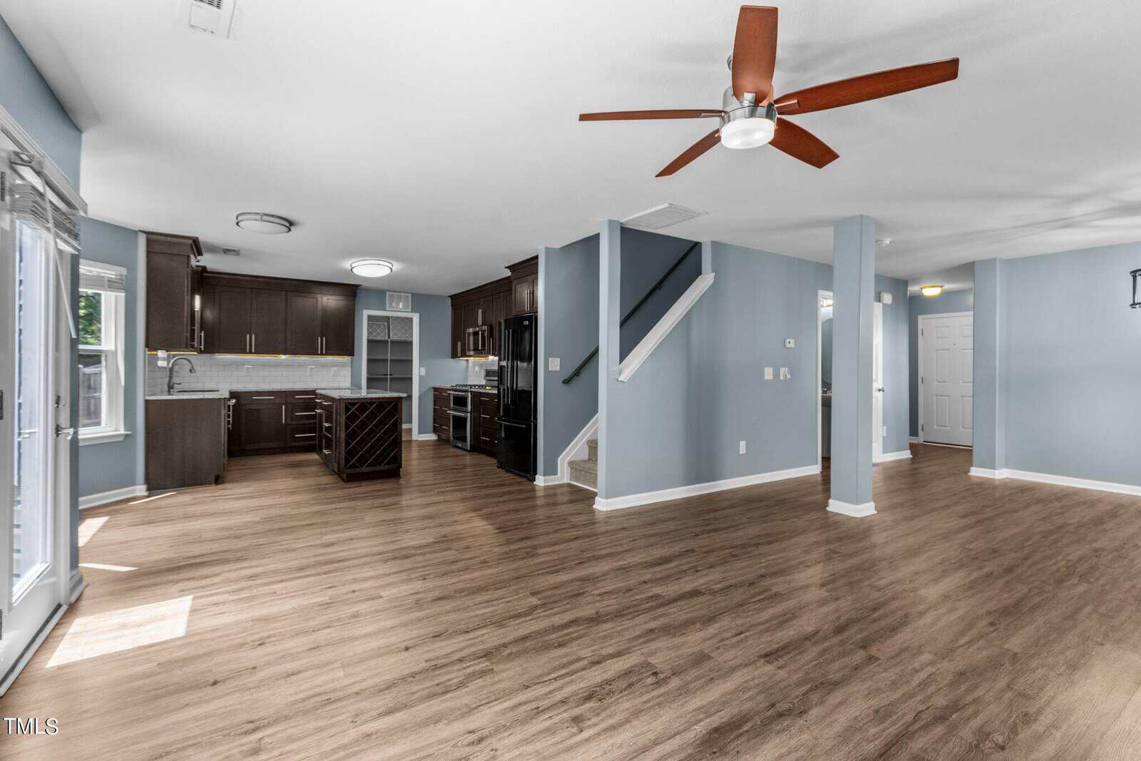 Spacious open-concept living area with light blue walls and wooden flooring. A kitchen with modern dark cabinets, marble countertops, and an island is on the left. A ceiling fan and stairs lead to the upper level. Natural light enters through large windows.