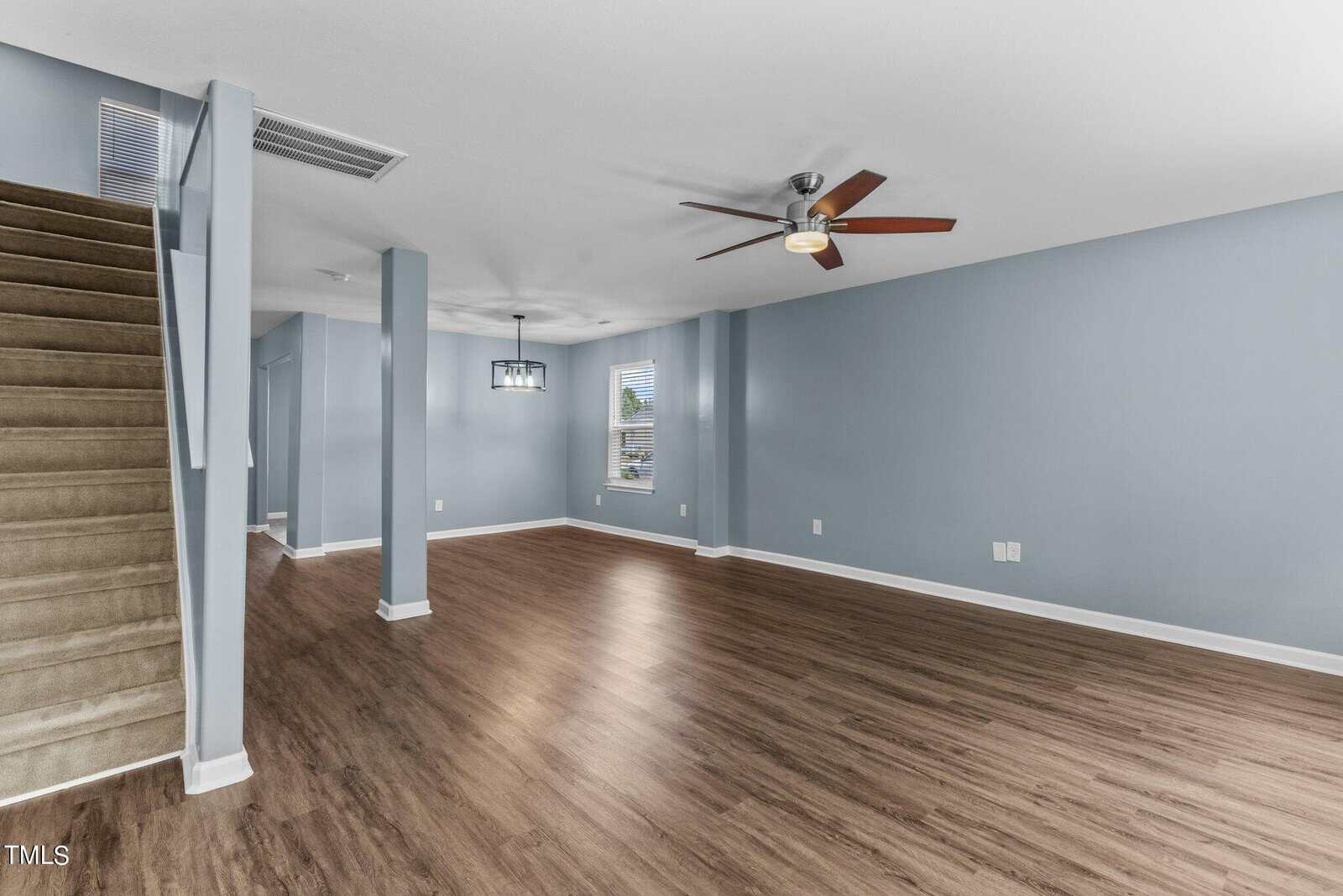 Spacious empty living room with light blue walls and wooden flooring. A ceiling fan with lights is centrally located. A staircase with beige carpeting is on the left, and a window provides natural light.