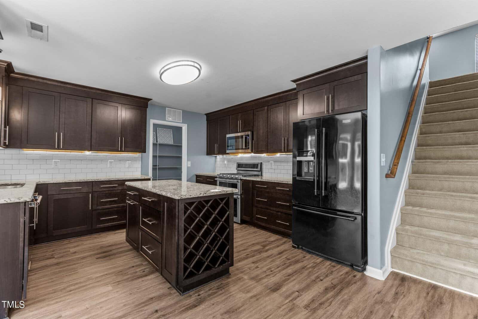Modern kitchen with dark wood cabinets, marble countertops, and a black refrigerator. A center island features built-in wine racks. The kitchen has stainless steel appliances, a light wood floor, and a nearby staircase with carpeted steps.