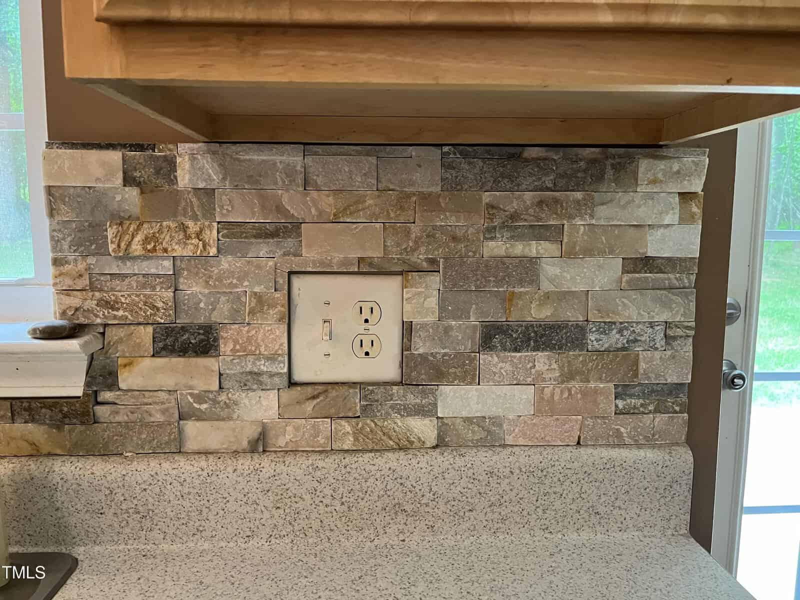 A kitchen backsplash features a mosaic of rectangular stone tiles in various shades of brown, grey, and beige. An electrical outlet is centered in the image, and a segment of a beige countertop is visible below.