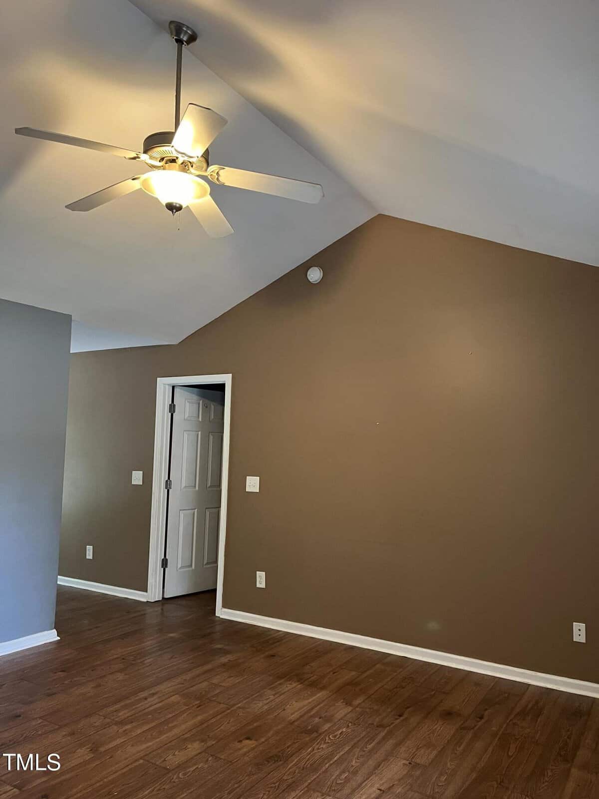 A room with a vaulted ceiling features a ceiling fan with a light. The walls are painted brown and gray, and the floor is hardwood. A white door leads to another room.
