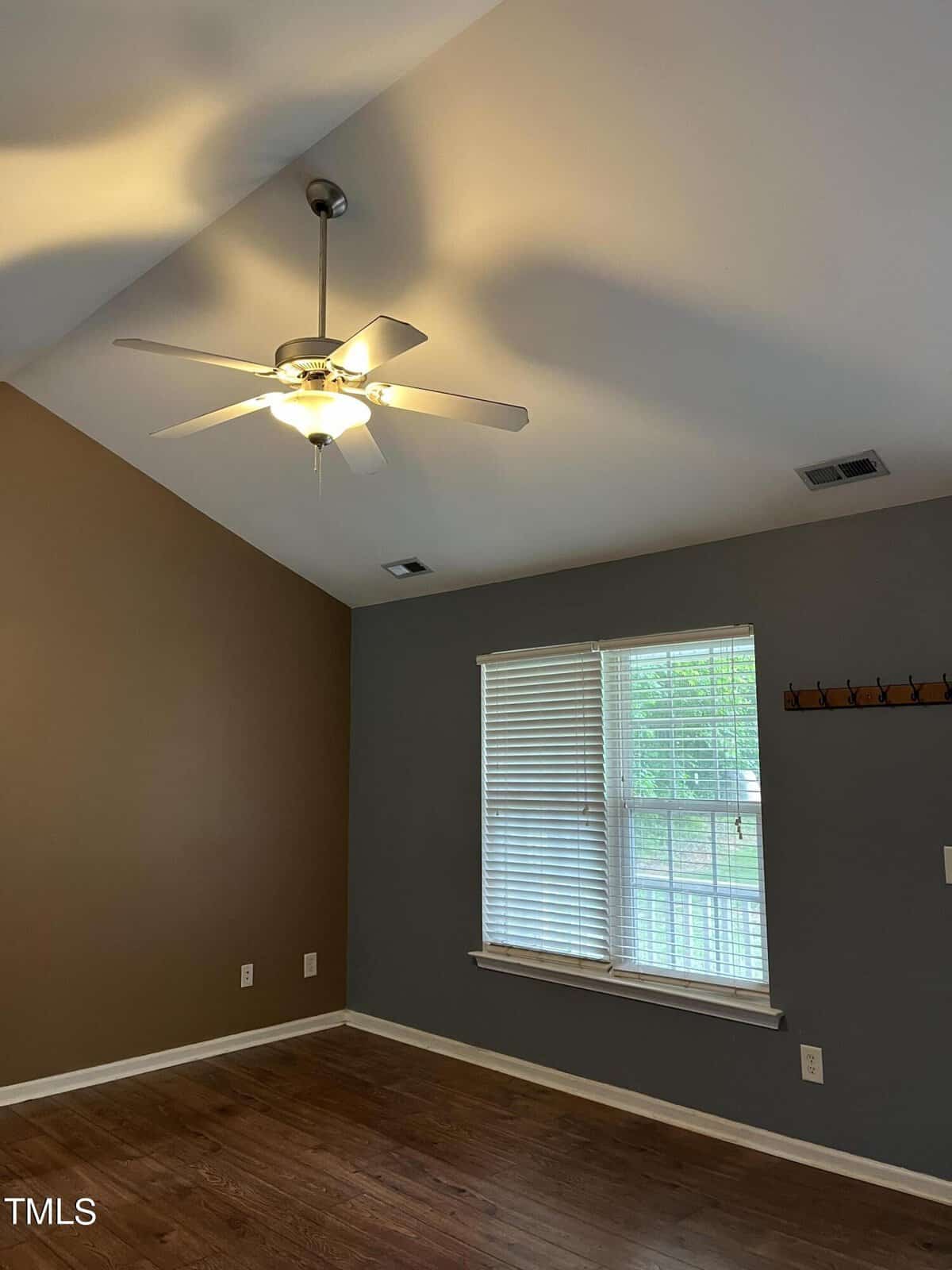 A room with a vaulted ceiling features a lit ceiling fan, two-tone walls in brown and gray, a three-panel window with blinds partly open, and a wooden floor. A coat rack is mounted on the gray wall.