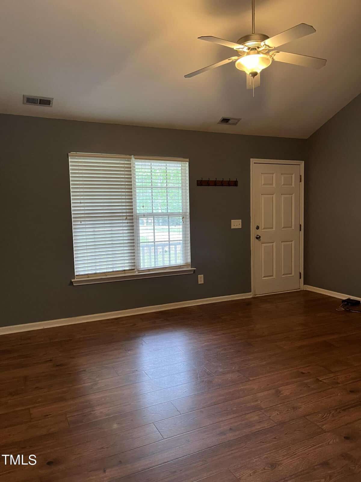 A room with wood flooring, a grayish-green painted wall, and a white ceiling fan. There is a window with blinds and a door on the adjacent wall. The room is empty, with some light coming in through the window.