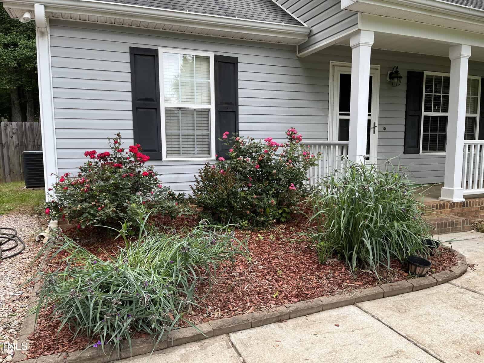 A small garden bed in front of a gray house with white trim features flowering shrubs with red blooms, green ornamental grasses, and mulch. A covered porch with white railing is partially visible. Black shutters frame the windows.