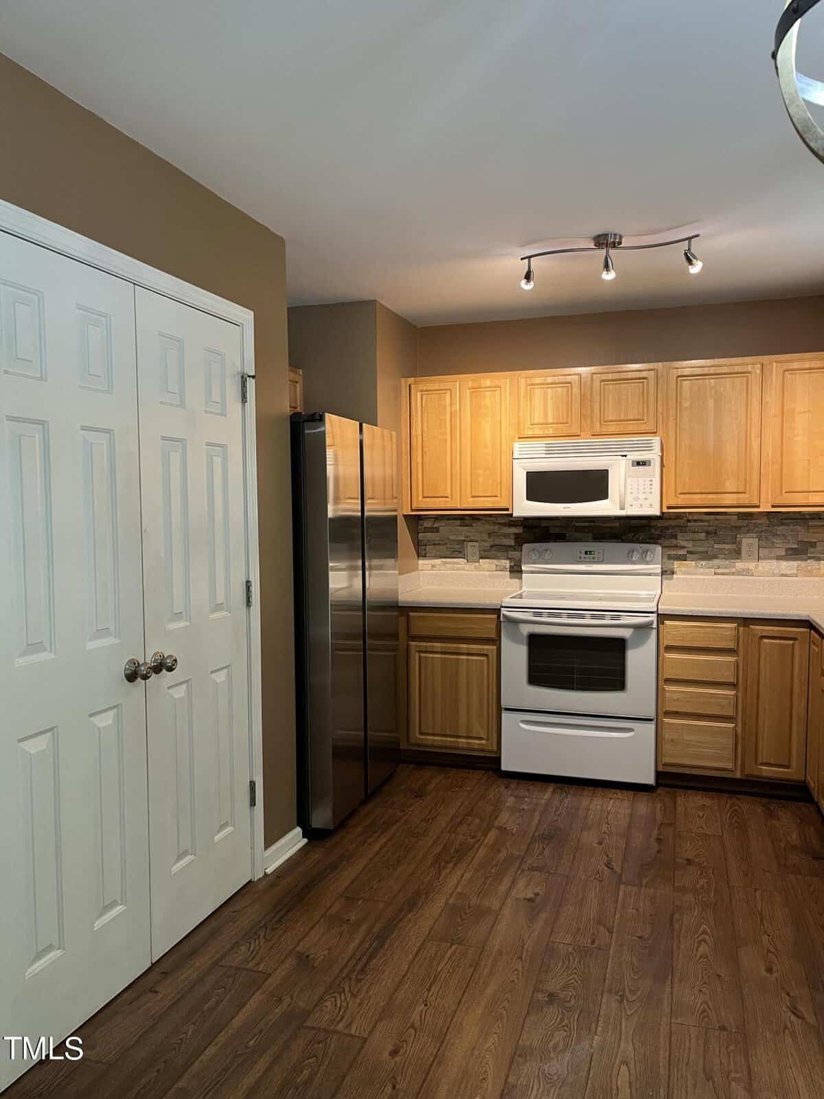 A modern kitchen with wooden cabinets, stainless steel fridge, and a white oven and microwave. The room features a tiled backsplash, hardwood flooring, and overhead track lighting. Double doors are visible to the left.