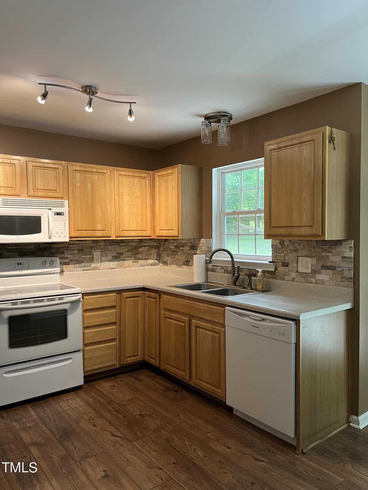 A kitchen with wooden cabinets, a white stove, microwave, and dishwasher. The countertop is light-colored, and the backsplash features a stone design. A window above the sink lets in natural light, and overhead lights are installed.