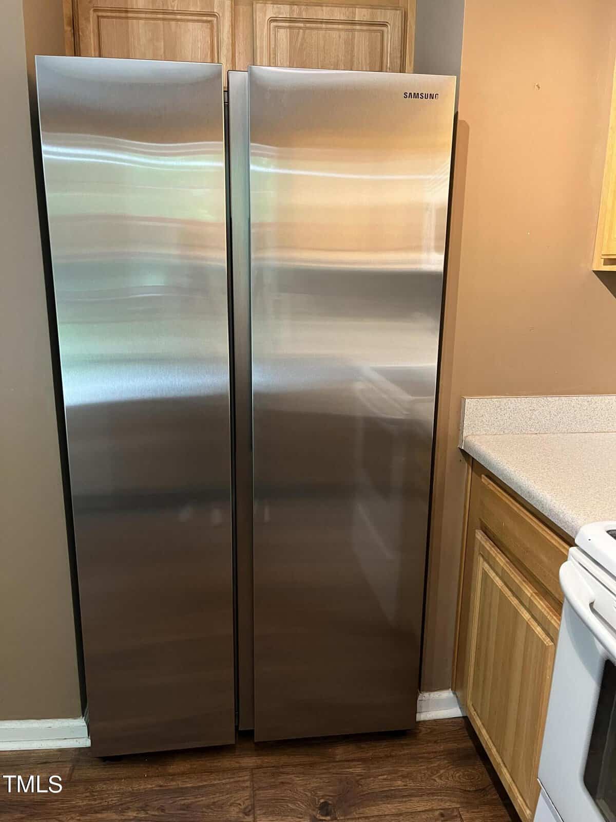 A sleek, stainless steel Samsung side-by-side refrigerator is situated between a wooden kitchen cabinet and a white stove, against a light brown wall. The fridge has a minimalist design and reflects the surrounding kitchen area.