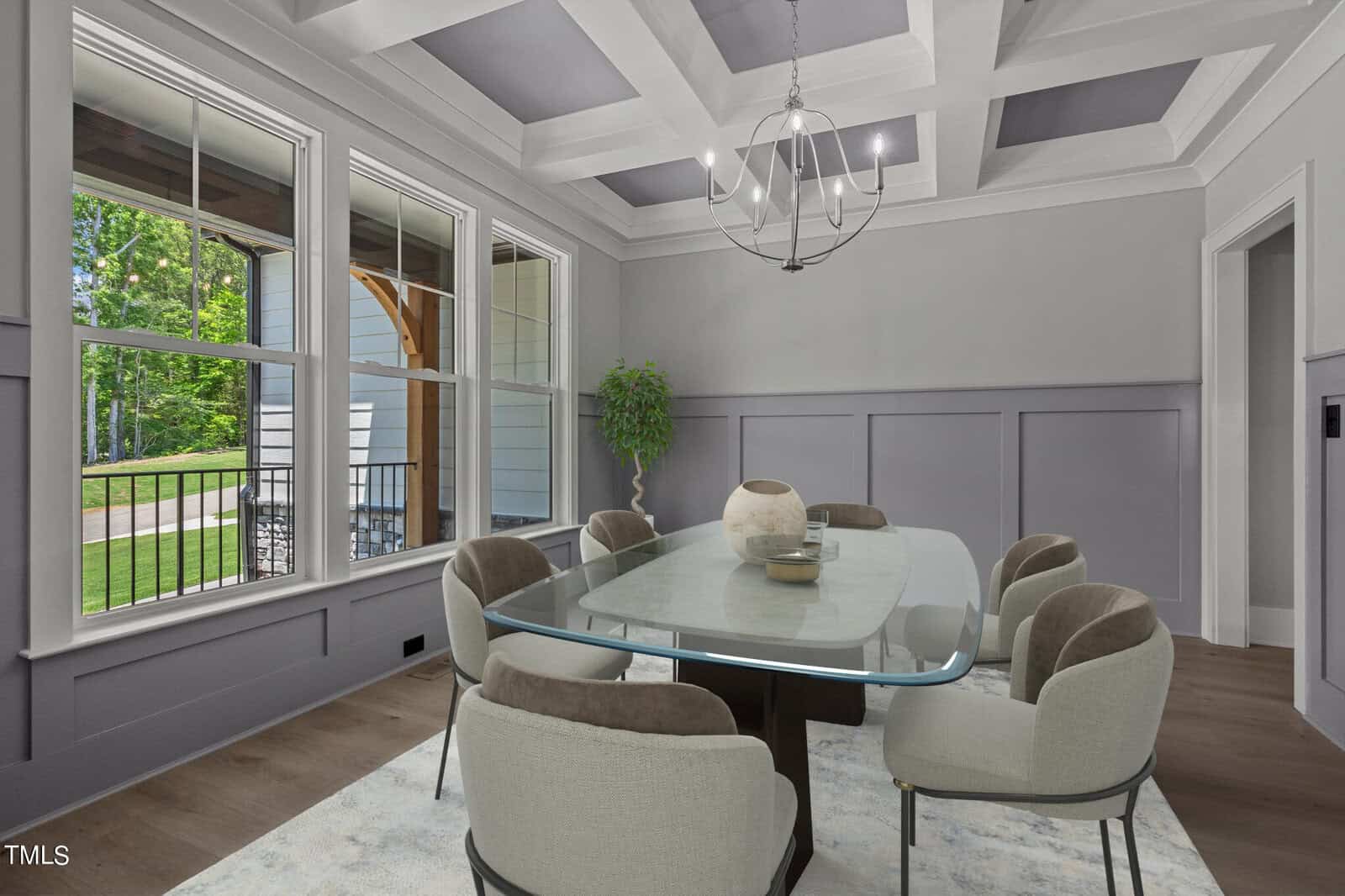 Elegant dining room with a coffered ceiling, featuring a glass-top table surrounded by upholstered chairs. A modern chandelier hangs above, and tall windows offer a view of the outdoors, letting in natural light. A small potted plant adds greenery.