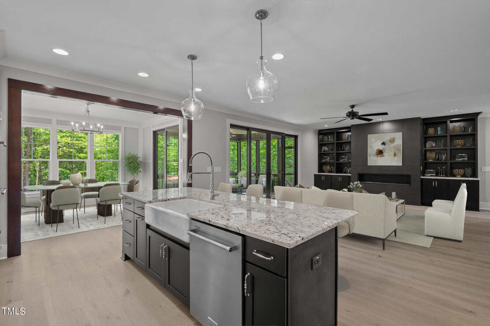 Spacious open-plan kitchen and living area with large windows and forest views. Features a kitchen island with a marble countertop, pendant lights, a dining table, and a comfortable seating area with a modern fireplace and built-in shelves.