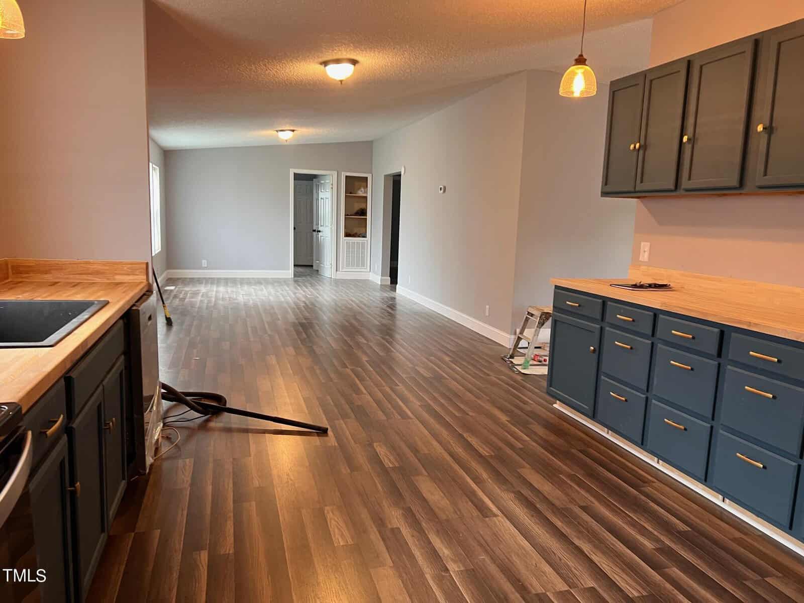 Spacious kitchen and living area with dark wood flooring, light gray walls, and dark blue cabinets. Recessed lighting and pendant lights provide illumination. A sink and countertop are visible, with a doorway leading to another room.