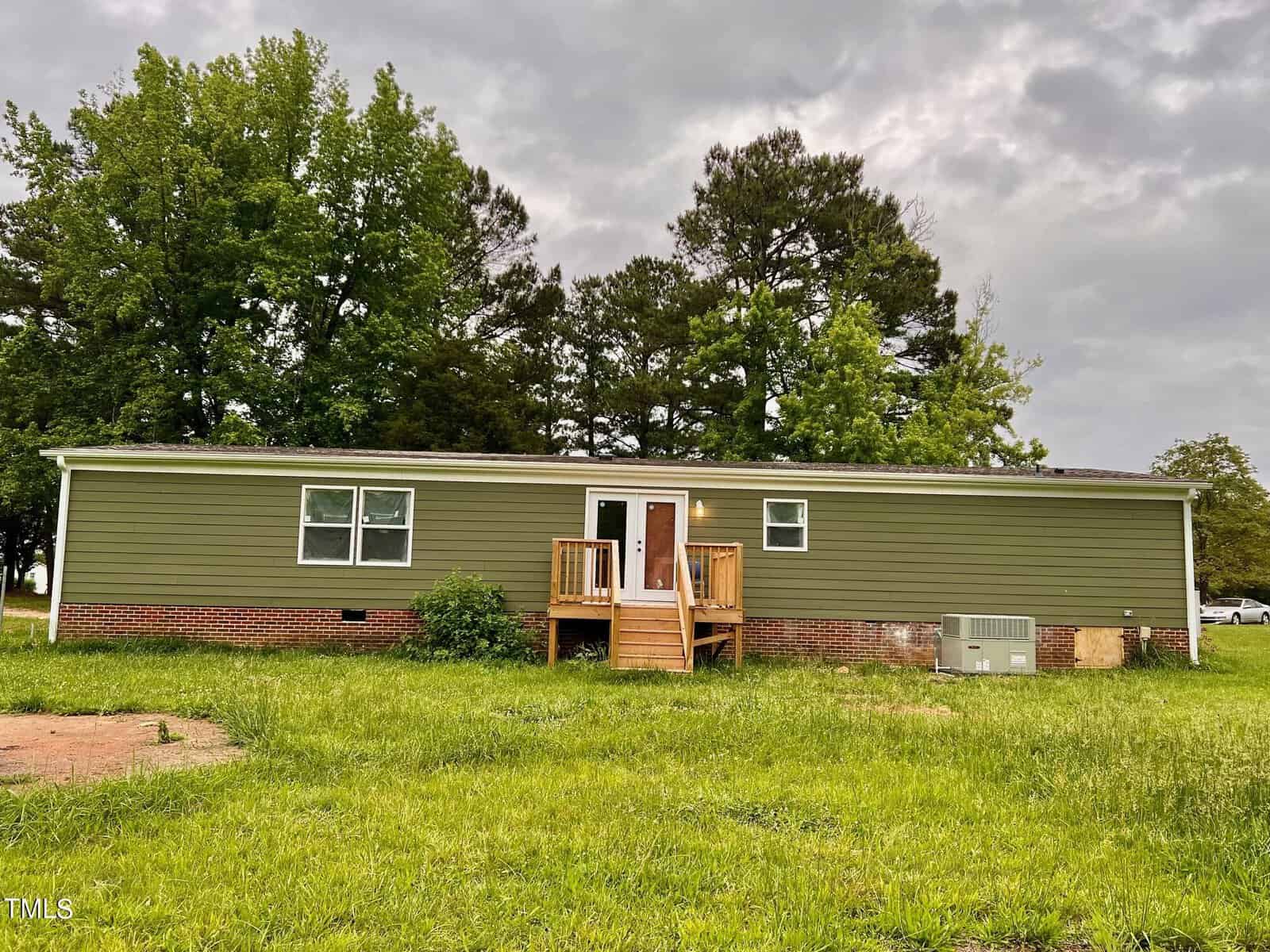A single-story green house with a small wooden porch and stairs, surrounded by a grassy area. Behind the house are tall trees, and the sky is overcast with gray clouds. A small air conditioning unit is visible on the side.