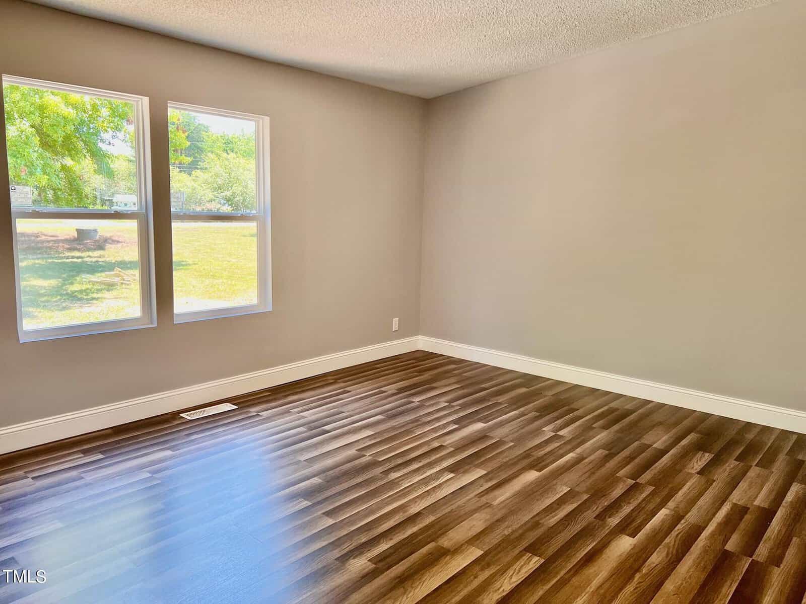 An empty room with light gray walls and dark wooden flooring. Two large windows on the left side let in natural light, showing a green outdoor view.