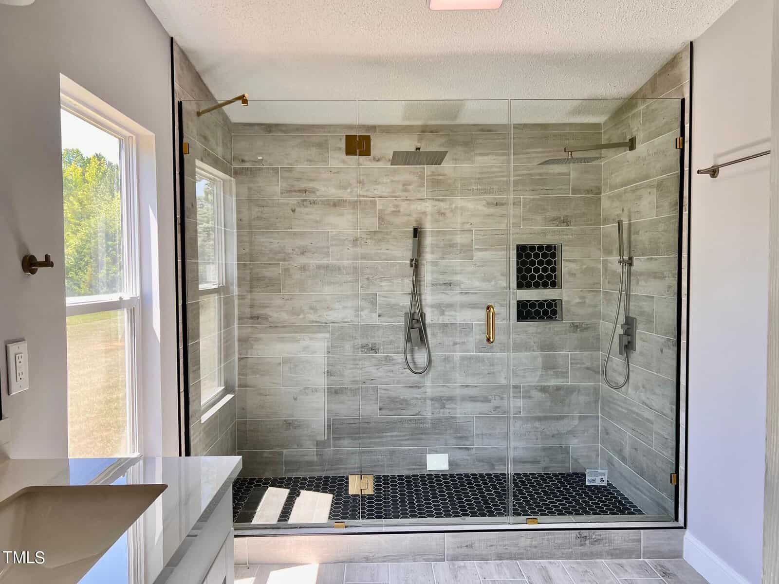 Modern bathroom with a large glass-enclosed shower. The shower features gray tile walls, dual rainfall showerheads, and a black hexagon tile floor with a built-in shelf. A window on the left lets in natural light.