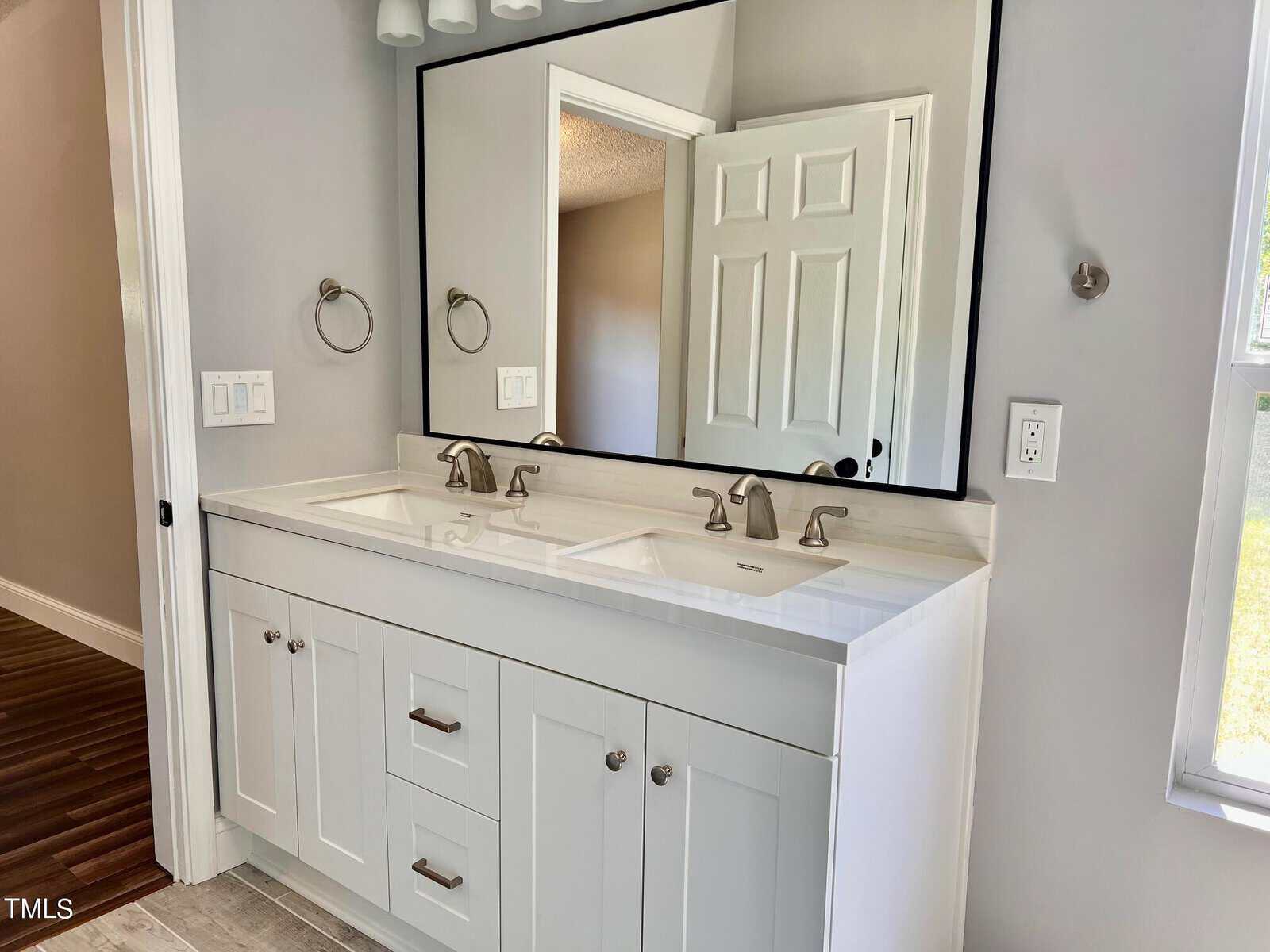 A modern bathroom features a double sink vanity with white cabinets and a marble countertop. Two square mirrors hang above the sinks, accompanied by wall-mounted light fixtures. The room is well-lit, with a window allowing natural light in.